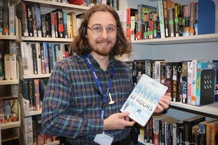 Librarian Jason holds up a book written by Lee Child, with a library bookshelf in the background