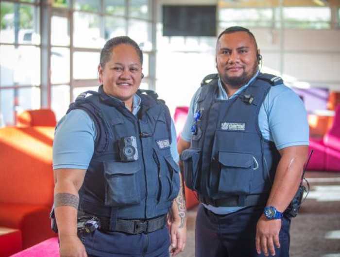 A pair of Corrections Officers from Auckland Region Womens Corrections Facility are picutred side by side, in a large open room with chairs in the background