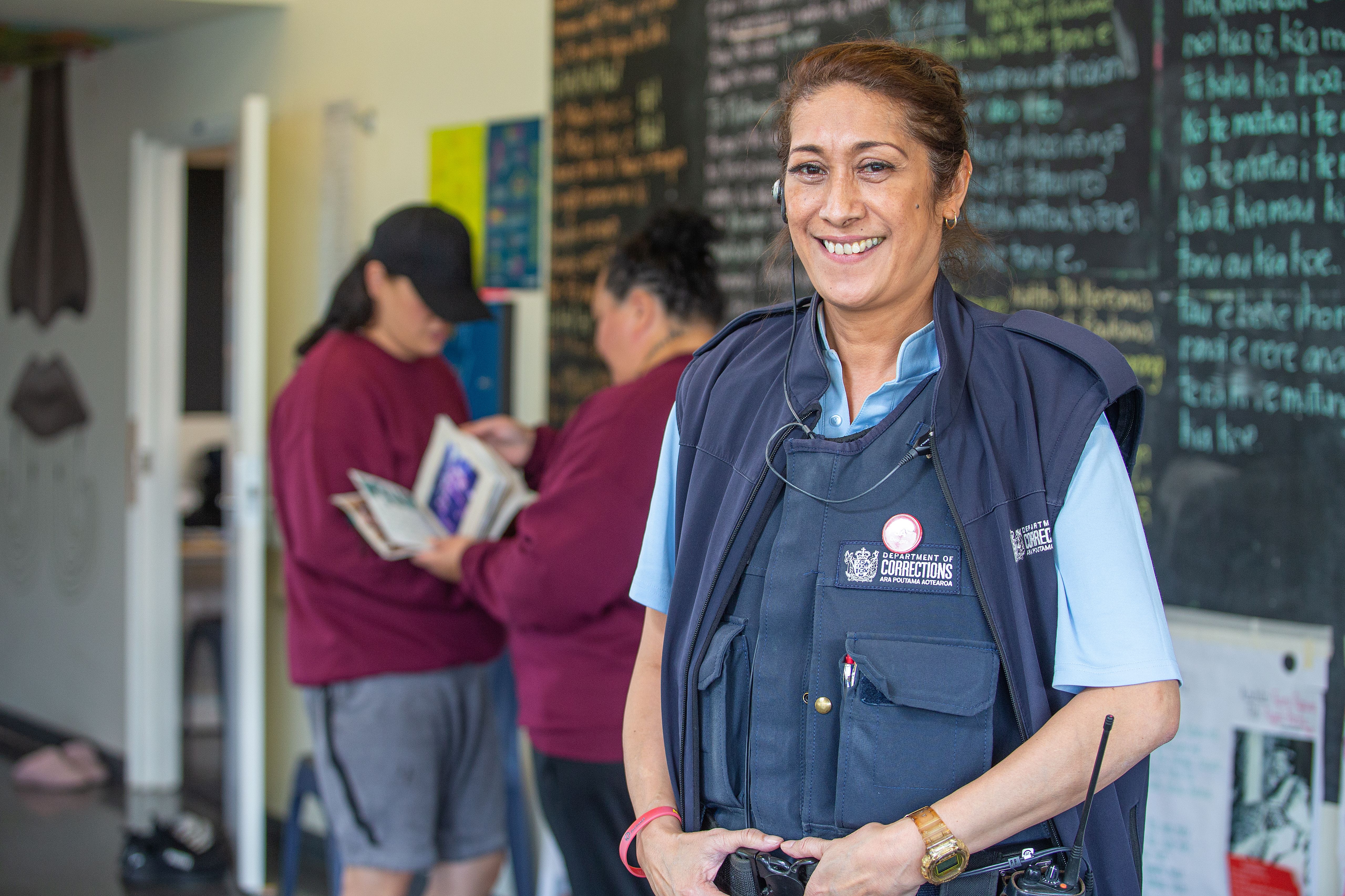 Corrections Officer stands smiling at camera. A blackboard with te reo Maori on is in the background.